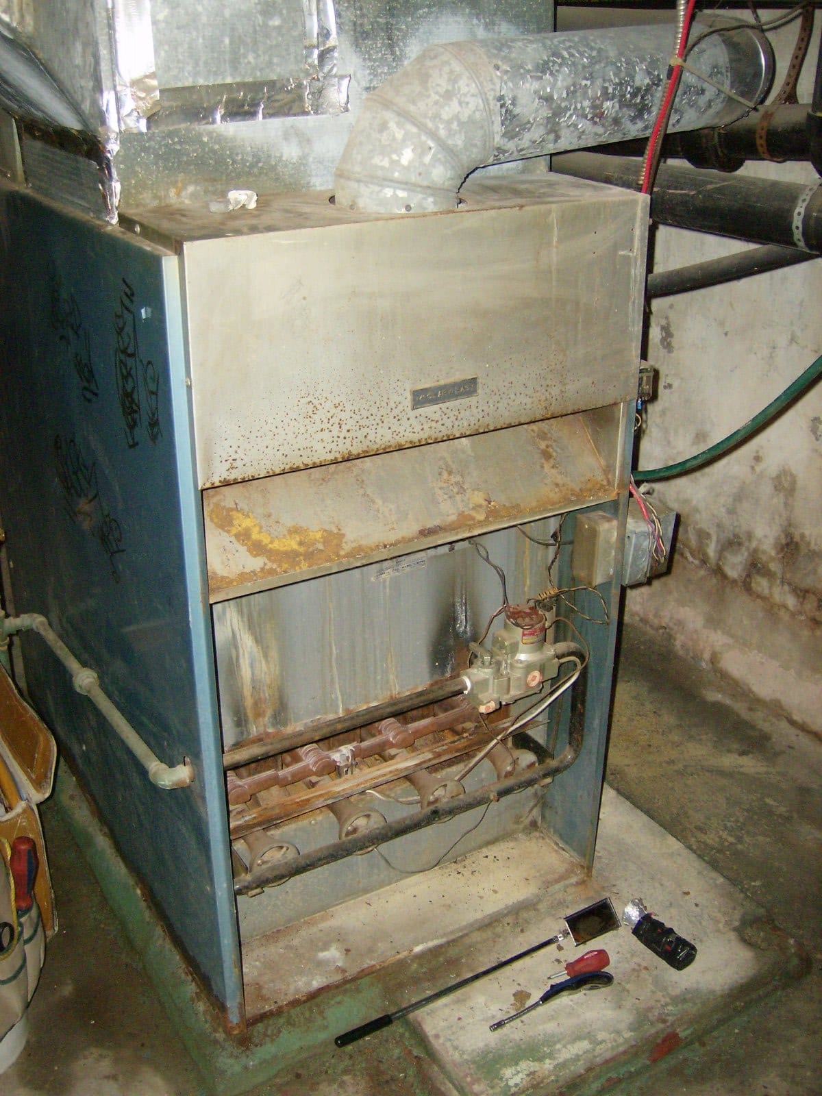 An older gas furnace in a basement with its front panel removed, showing the row of burners and the gas valve