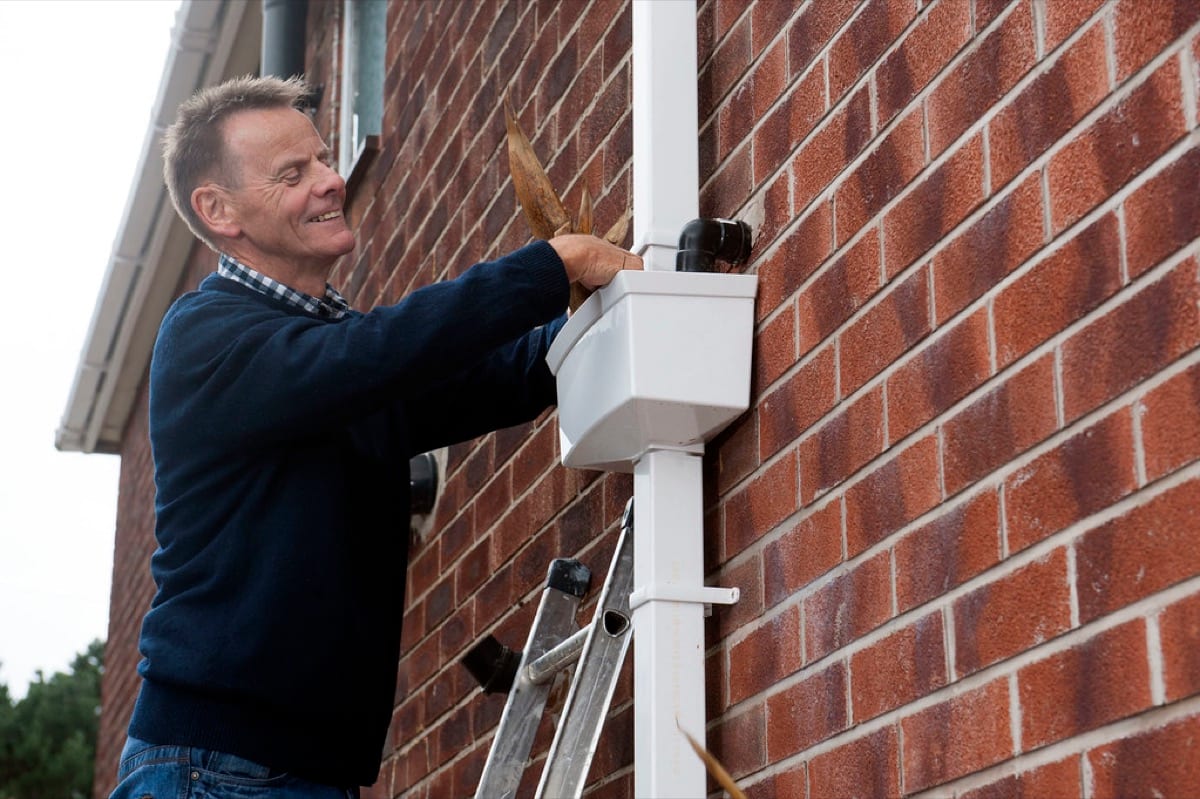 A person on a ladder pulling debris out of a gutter hopper on a brick house