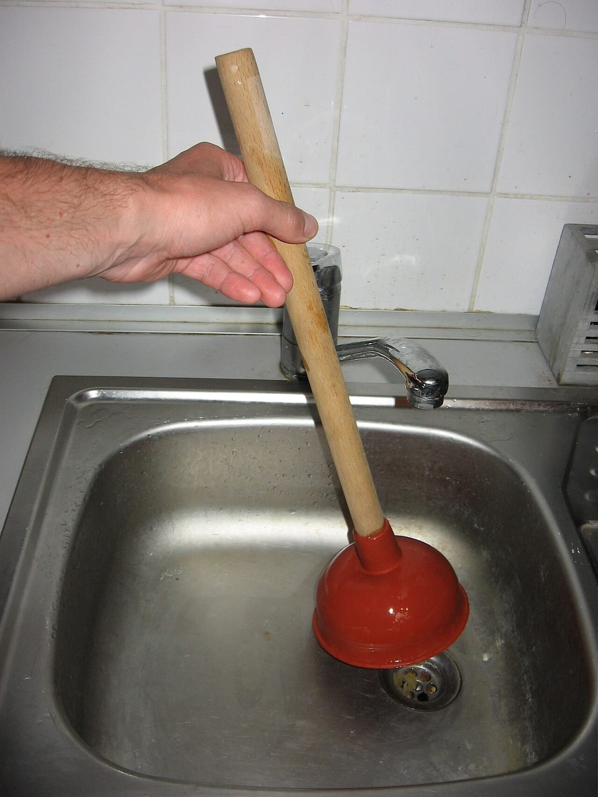 A hand working a red cup plunger over the drain of a stainless steel kitchen sink