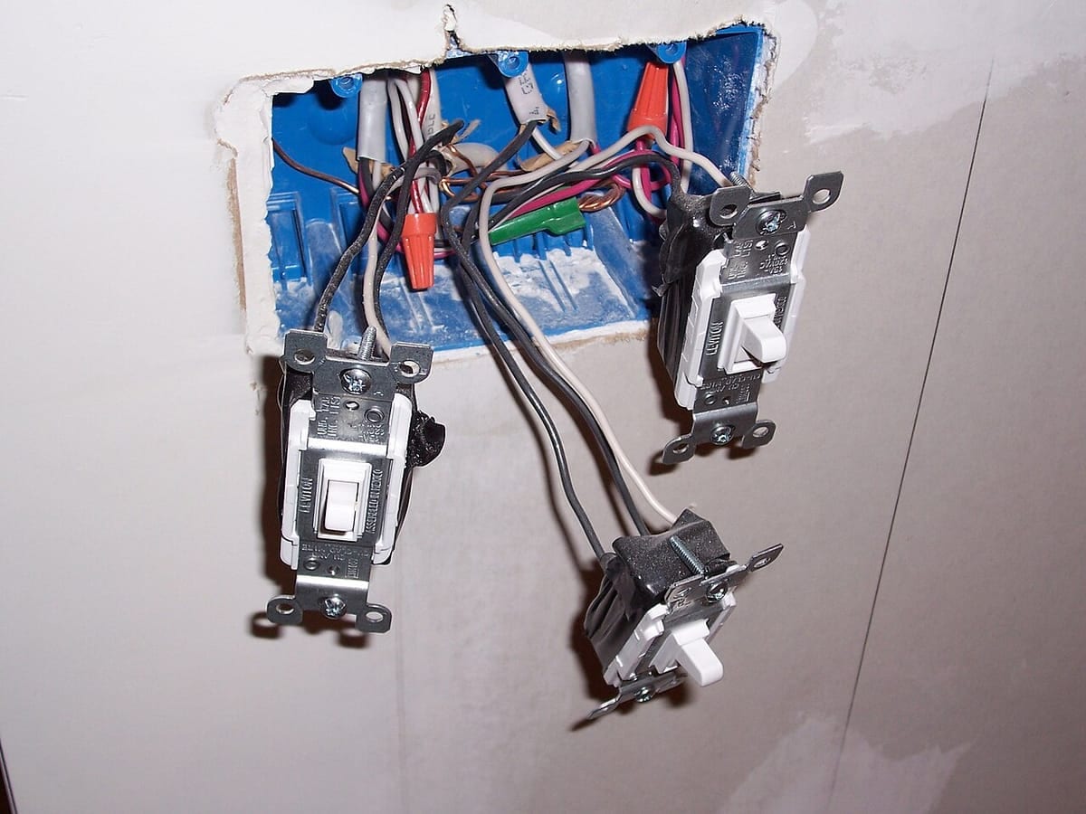 Three toggle light switches pulled out of a blue plastic wall box, hanging by their wires