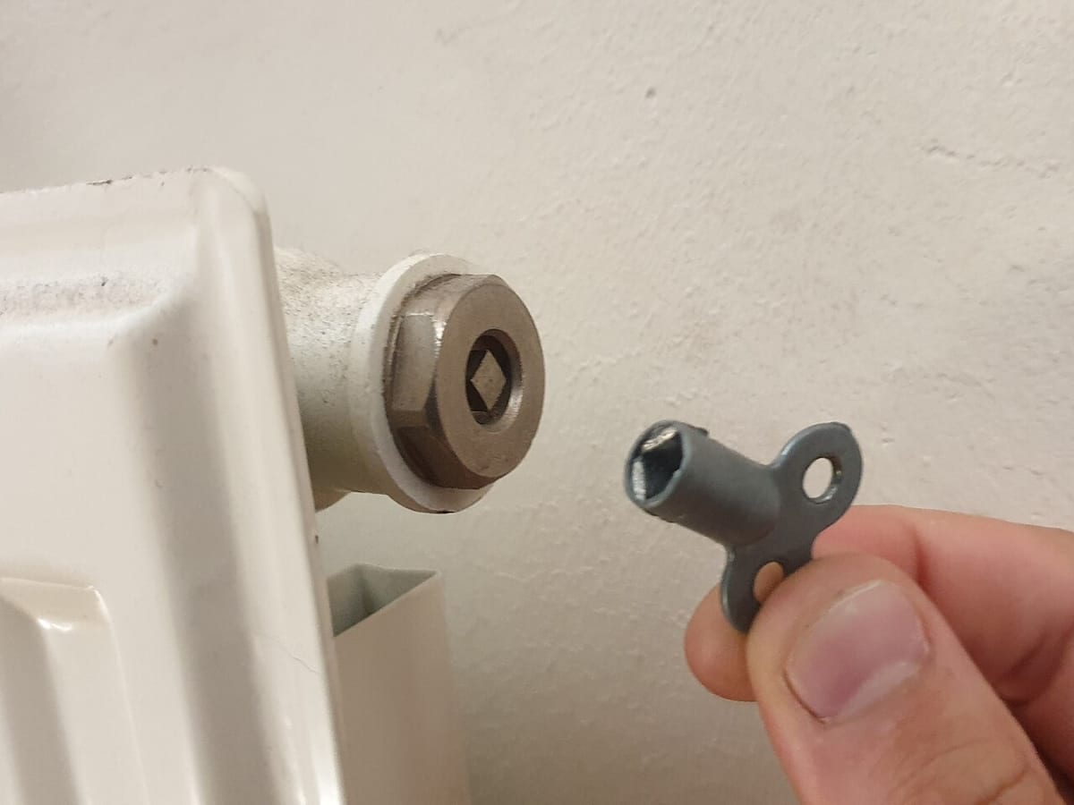 A hand holding a radiator key beside the square bleed valve on a white radiator