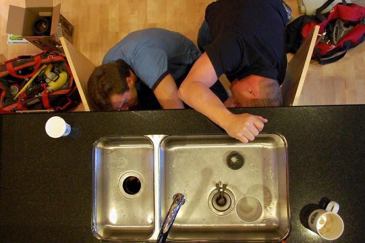 Two people working underneath a kitchen sink, seen from above through the sink basins